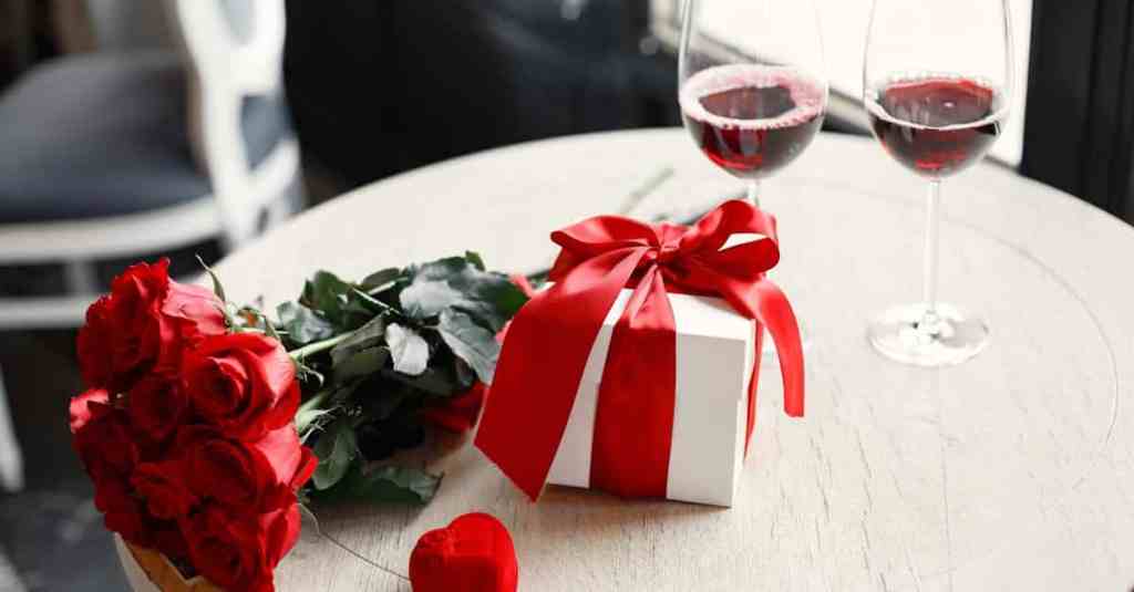 A romantic table setting with red roses, gift box, and red wine glasses, perfect for Valentine's Day celebration.